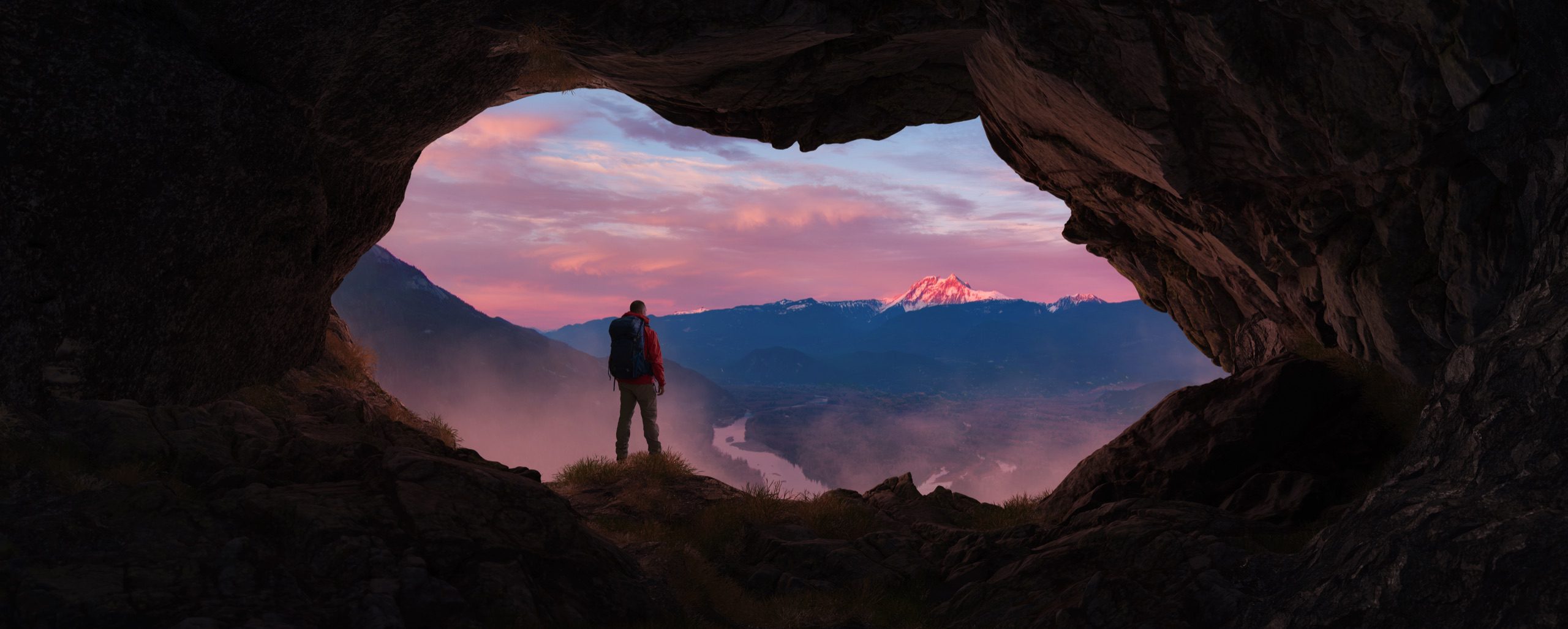 Escursionista all'imbocco di una grotta davanti a un panorama di montagne al tramonto